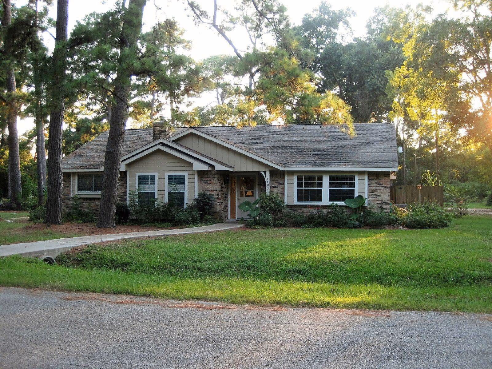Single-story house with a sloped roof, beige siding, and brick accents, surrounded by trees and greenery with a paved walkway leading to the front door.