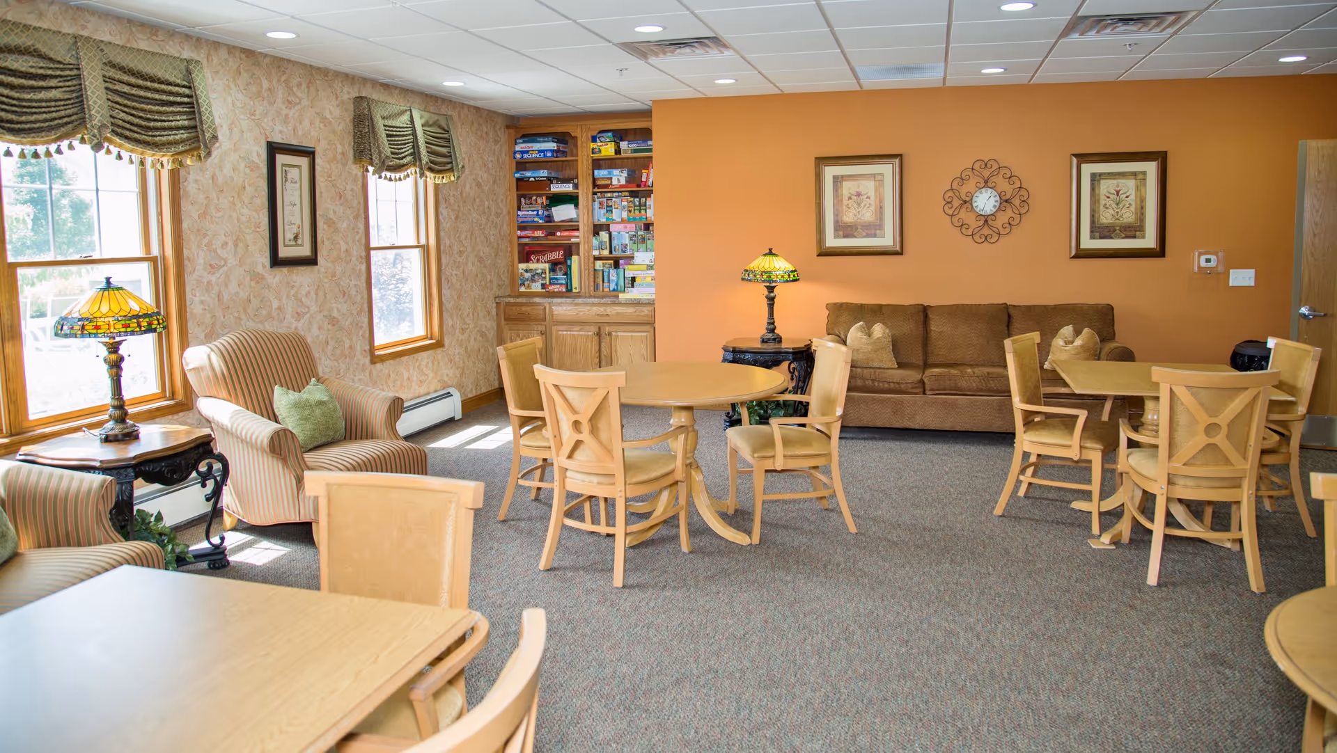A cozy senior living community room with multiple wooden tables and chairs, a brown couch against an orange wall, two framed pictures and a decorative clock above the couch. There are two armchairs with striped upholstery near windows with patterned valances, a side table with a Tiffany-style lamp, and a built-in wooden bookshelf filled with board games.