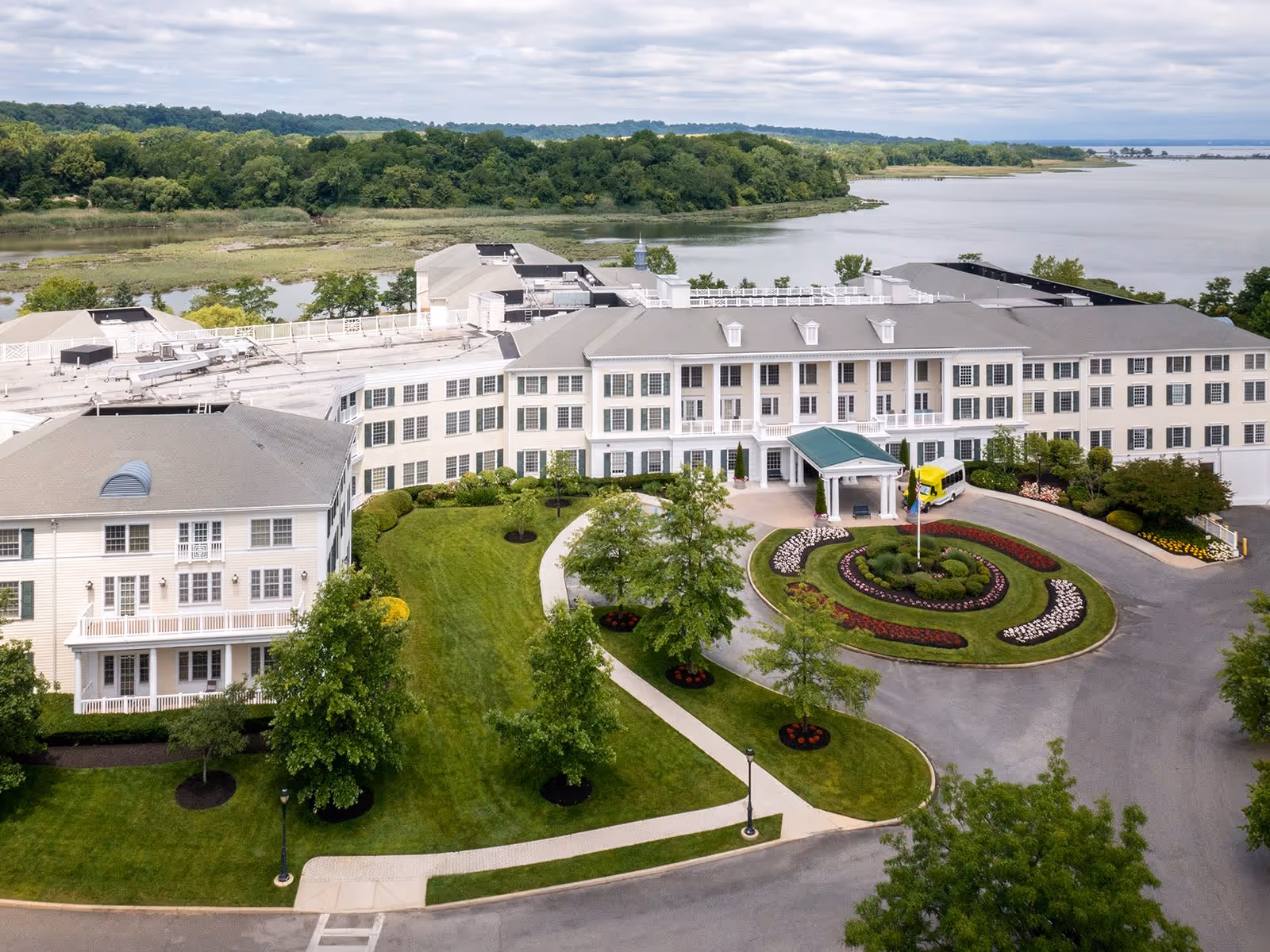 A large senior living facility named Atria on Roslyn Harbor with a circular driveway and landscaped garden in front, surrounded by green lawns and trees, located near a body of water with a forested area in the background.