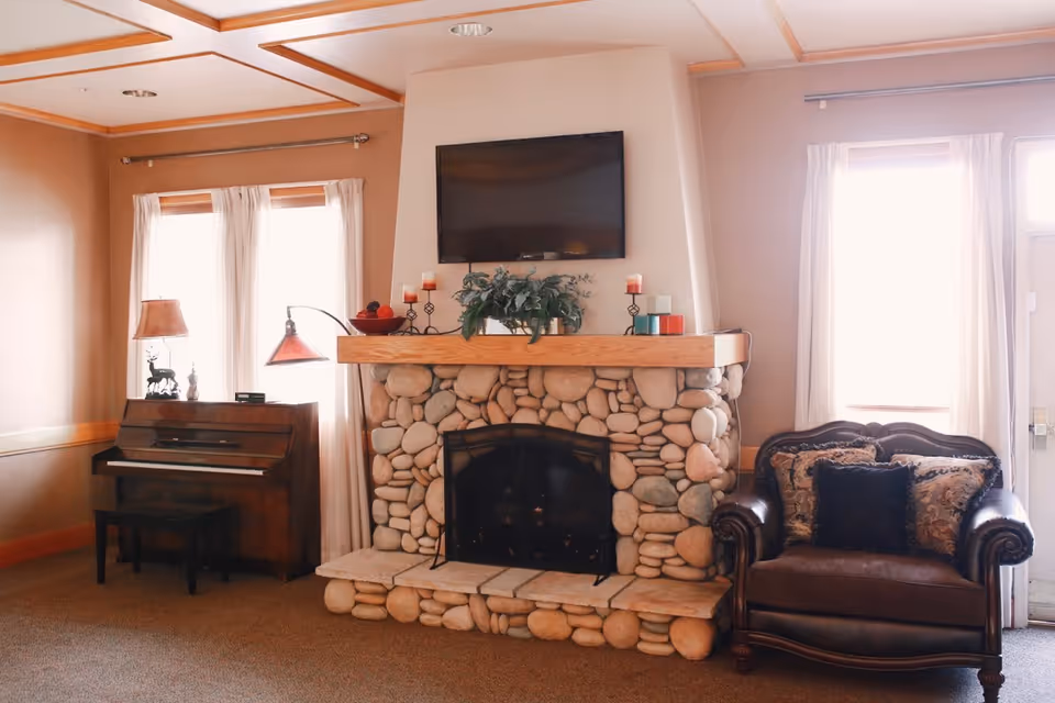 Cozy living room area with a stone fireplace in the center, a flat-screen TV mounted above it, and decorative candles and plants on the mantel. To the left of the fireplace is a wooden piano with a lamp and decorative items on top. To the right is a dark leather armchair with patterned cushions. The room has beige walls, carpeted floor, and windows with white curtains letting in natural light.