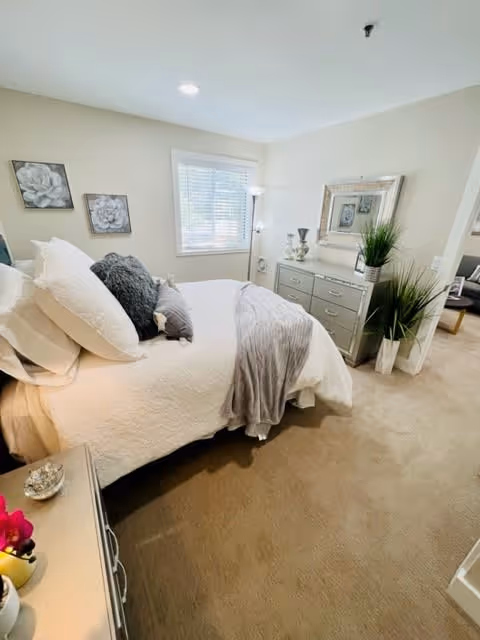 A cozy bedroom with a neatly made bed featuring white and gray bedding. The room has beige walls and carpet, a window with blinds, two framed floral artworks on the wall, a dresser with decorative items and a large mirror above it, and a tall floor lamp in the corner. There is an open doorway leading to another room with a chair and table visible.