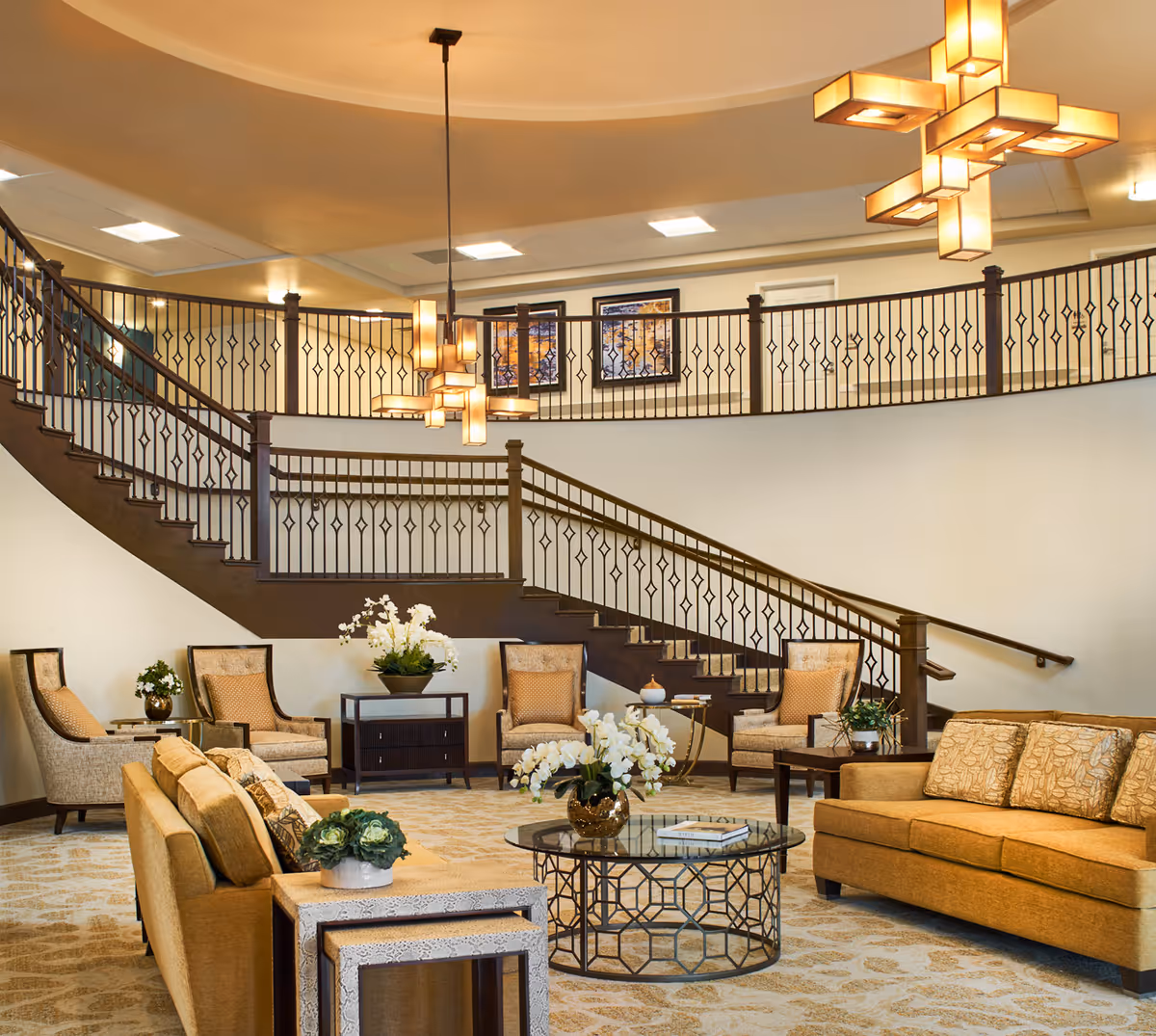 A spacious and elegant living room area with beige and gold-toned sofas and armchairs arranged around a glass coffee table with a decorative plant. The room features a curved staircase with dark wooden railings and two modern pendant lights hanging from the ceiling. The walls are light-colored with framed artwork displayed on the upper level.