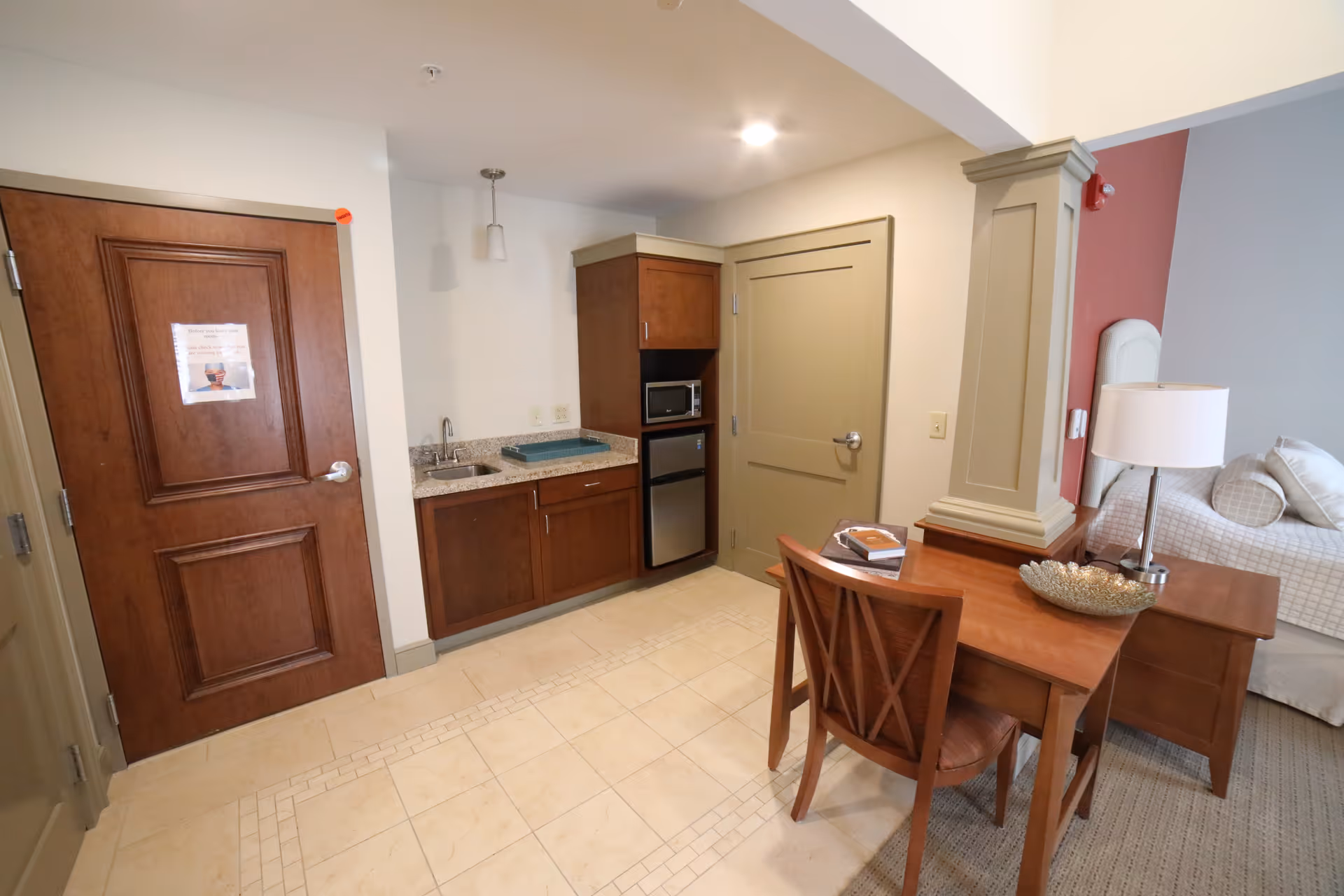 Interior view of a senior living facility room showing a small kitchenette with a sink, microwave, and mini refrigerator. There is a wooden table with a chair, a lamp, and decorative bowl. A bed with pillows is partially visible in the background. The room has tiled flooring near the kitchenette and carpet near the bed area.
