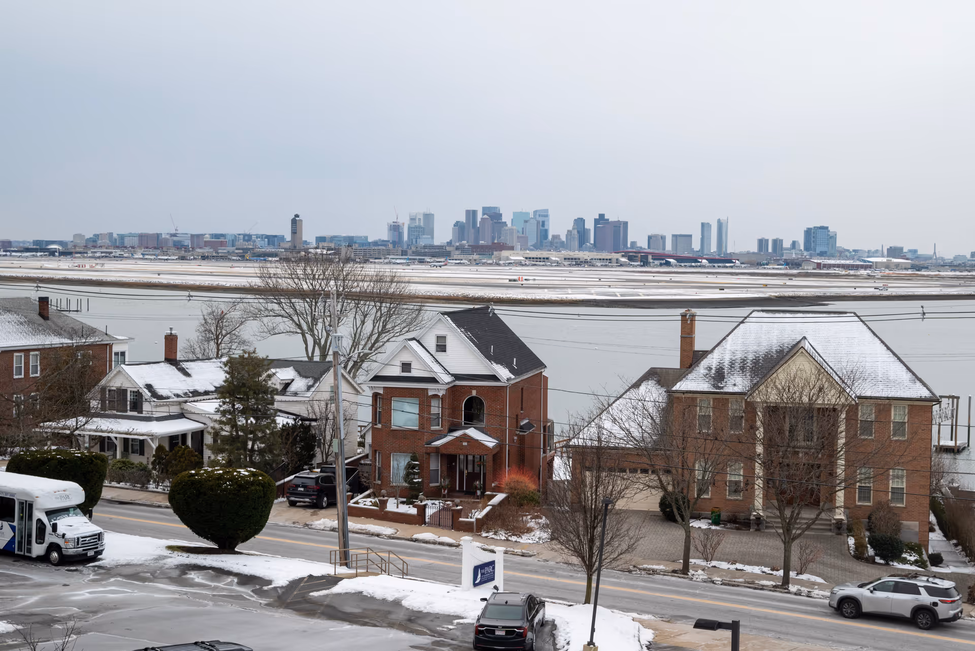 View of a snowy suburban neighborhood with brick houses and a road in the foreground, a body of water behind the houses, and a city skyline in the distance under a cloudy sky.