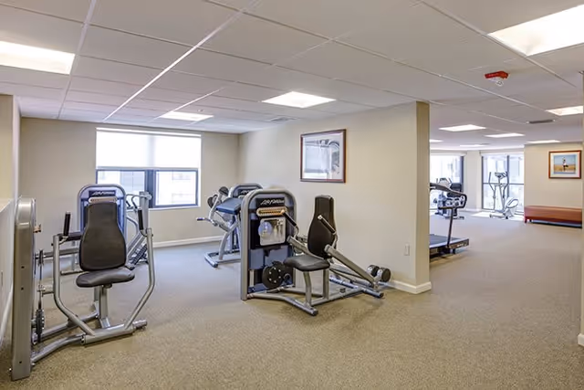 A fitness room in a senior living facility with exercise machines including a seated leg press and other strength training equipment. The room has beige carpet, white walls, a drop ceiling with fluorescent lights, and windows letting in natural light. There are framed pictures on the walls and an open area leading to more exercise equipment in the background.
