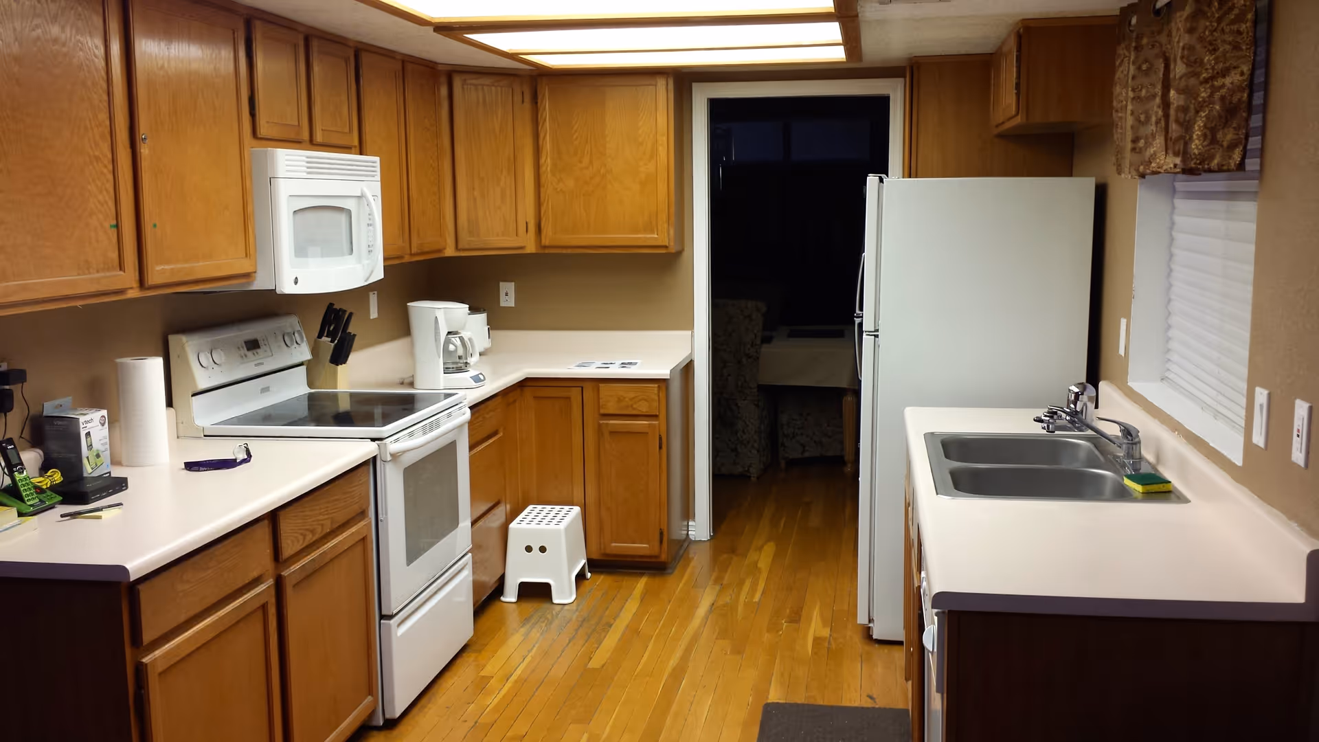 Interior view of a kitchen with wooden cabinets, white countertops, a white stove with an overhead microwave, a coffee maker, a white refrigerator, a double sink, and a small white step stool on a wooden floor. There is a window with a patterned valance above the sink and a doorway leading to another room with furniture visible.