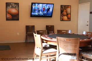 A room with a round poker table surrounded by wooden chairs. On the wall, there is a flat-screen TV mounted above two framed pictures of basketballs. The floor is wooden, and there is a door on the right side of the room.