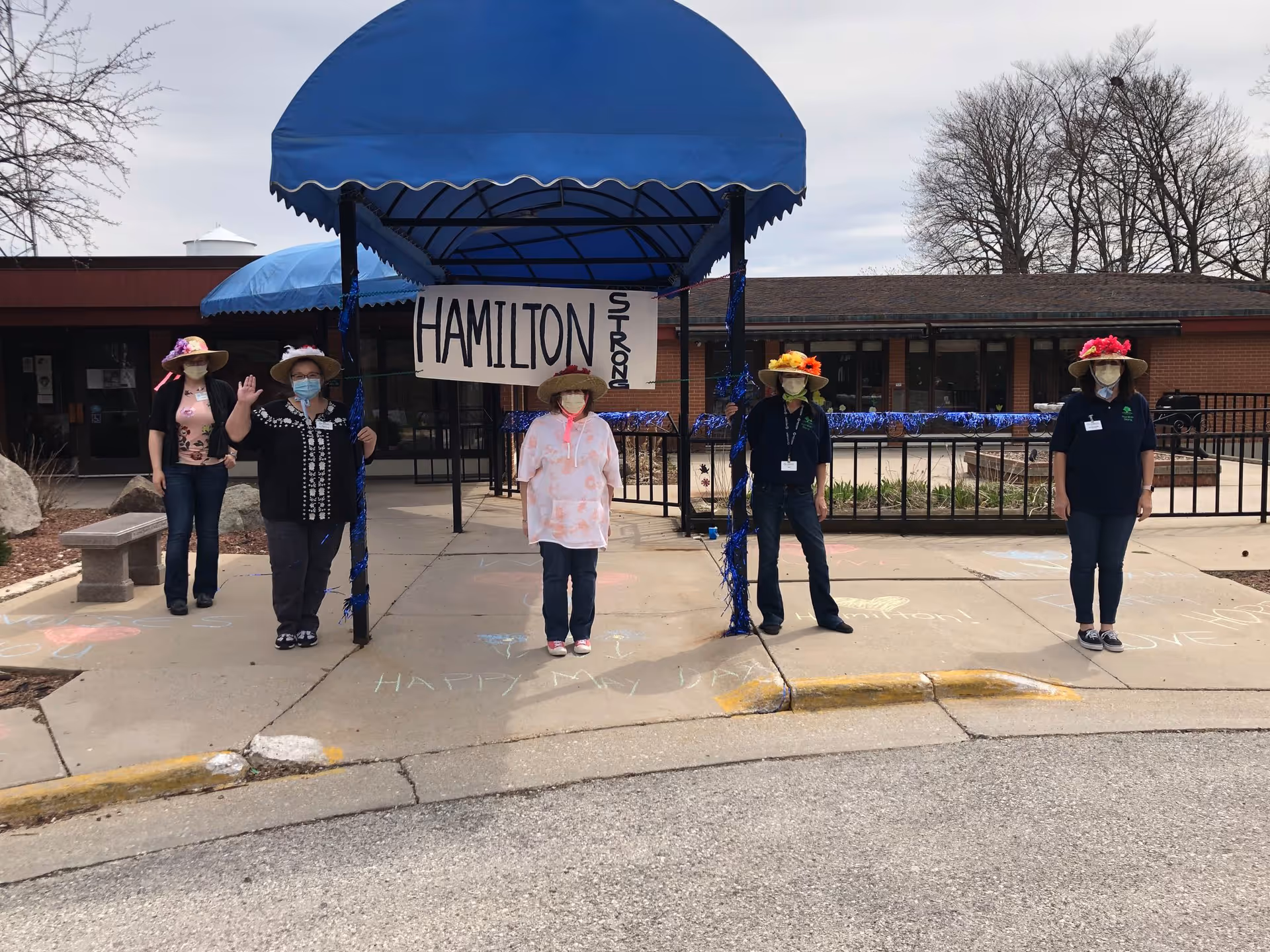 Five staff wearing face masks and decorative hats stand spaced under a blue awning outside the Hamilton Health Services entrance with a sign reading "HAMILTON".