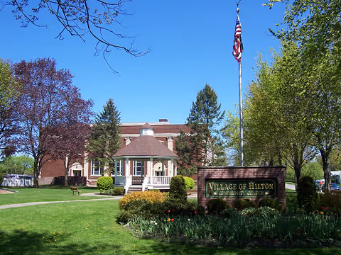 Exterior view of the Village of Hilton Assisted Living facility showing a brick building with a small gazebo-like structure in front, surrounded by green lawns, trees, and a flagpole with an American flag.