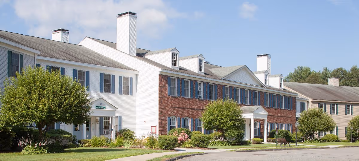Exterior view of a senior living facility building with white and red brick walls, multiple windows with blue shutters, and several chimneys. The building is surrounded by green bushes, trees, and a well-maintained lawn under a clear blue sky.