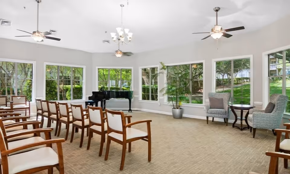 A bright room with large windows showing a green outdoor view. The room has multiple wooden chairs with white cushions arranged in rows facing a black grand piano. There are two patterned armchairs with a small table between them near the windows. Ceiling fans and a chandelier provide lighting, and a potted plant adds greenery inside.