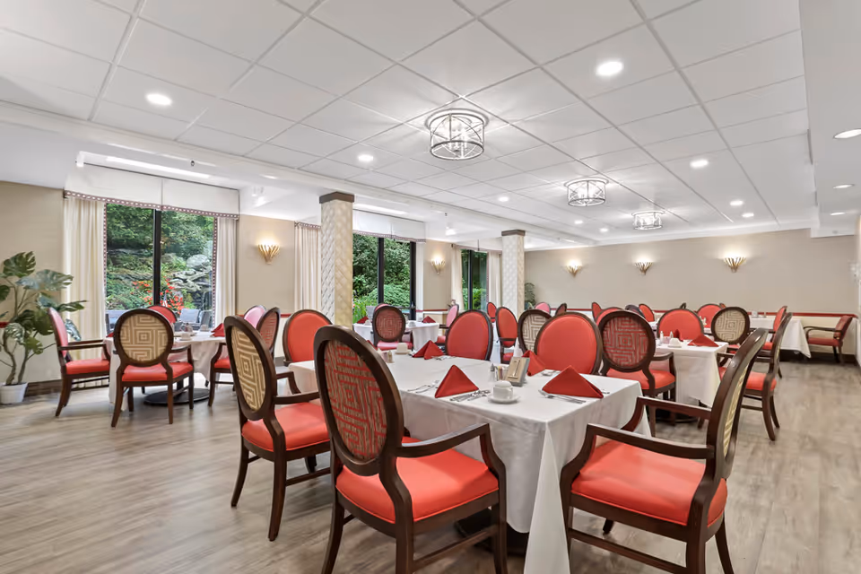 A spacious dining room with multiple tables covered in white tablecloths, each set with red folded napkins, cups, and silverware. The chairs have wooden frames with red and patterned upholstery. Large windows along one wall provide a view of greenery outside. The room has a light-colored ceiling with recessed lighting and decorative ceiling fixtures, and the floor is wood.