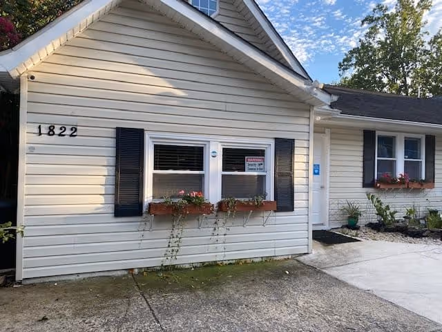 Exterior view of a single-story residential building with beige siding, black shutters, and flower boxes under the windows. The house number 1822 is visible on the left side. There is a concrete driveway and a small garden area near the entrance door.