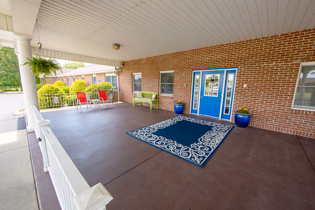 Covered entrance area of a senior living facility with a blue double door, a decorative blue and white rug, two blue flower pots with plants, a green bench, and two red chairs with a small table. The area has a white ceiling and brick walls, with greenery visible outside.