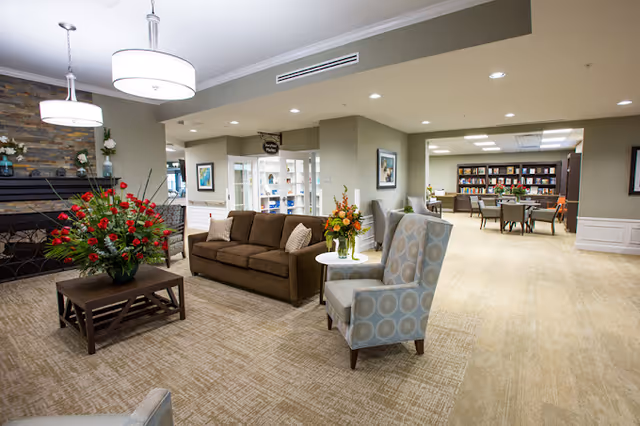 Bright communal lounge with sofas, armchairs, floral arrangements, and a view into a library/activity area.