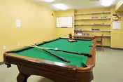 A recreation room with a green felt pool table in the center. Two pool cues rest on the table along with a rack of billiard balls. The room has beige walls, a whiteboard on the far wall, shelves with various items, and a wooden chair.