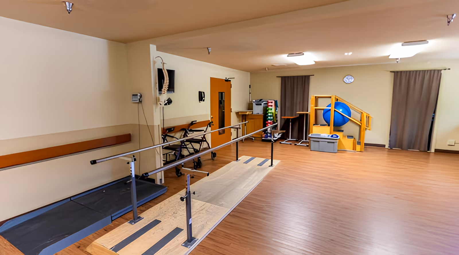 Spacious rehabilitation therapy room with parallel walking bars, walkers, exercise equipment, and a small therapy staircase with a blue exercise ball.
