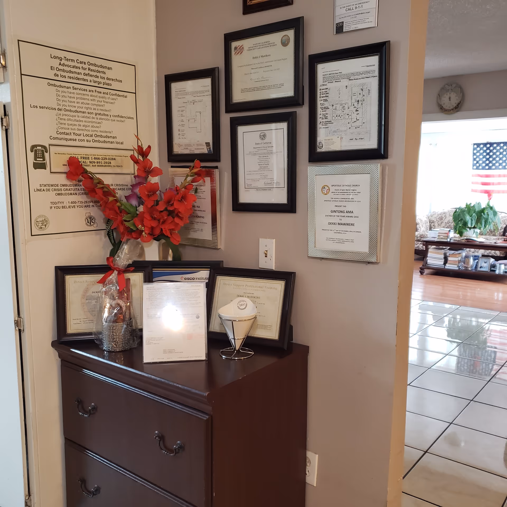 Interior corner showing a dark wooden chest with framed certificates and a vase of red flowers beneath wall-mounted diplomas, with a tiled room and an American flag visible through a doorway.