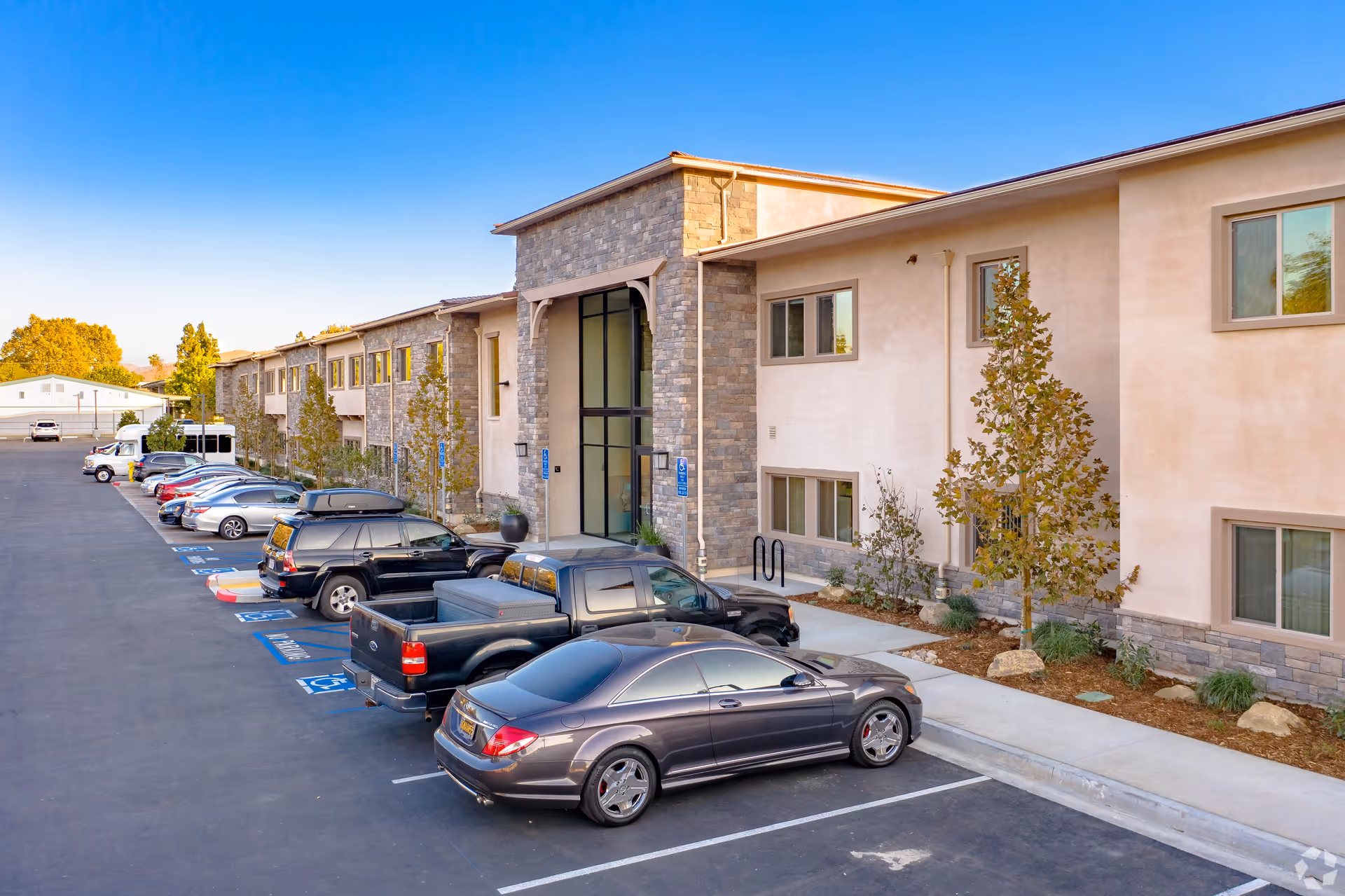 Exterior view of a two-story senior living facility building with stone and beige stucco facade, large glass entrance, and a parking lot with several parked cars including handicapped spaces.