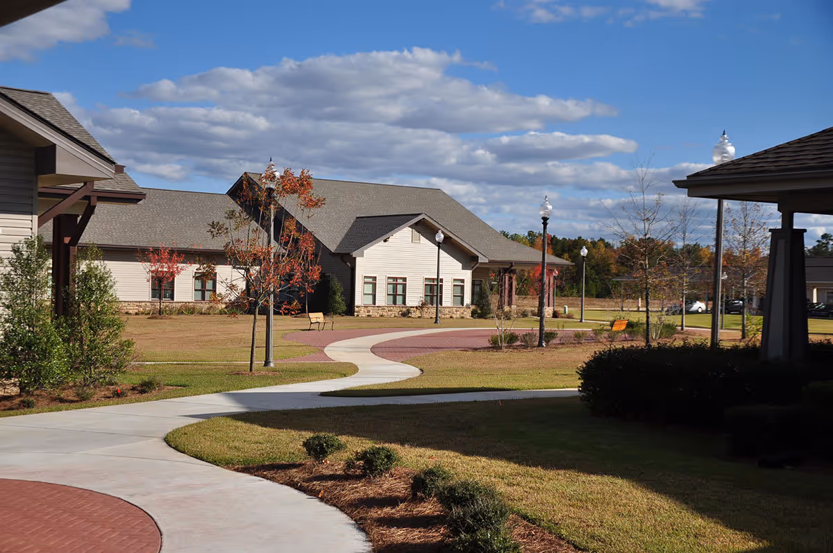 Exterior view of a senior living campus with a winding sidewalk, lawns, lamp posts, and single-story buildings under a partly cloudy sky.
