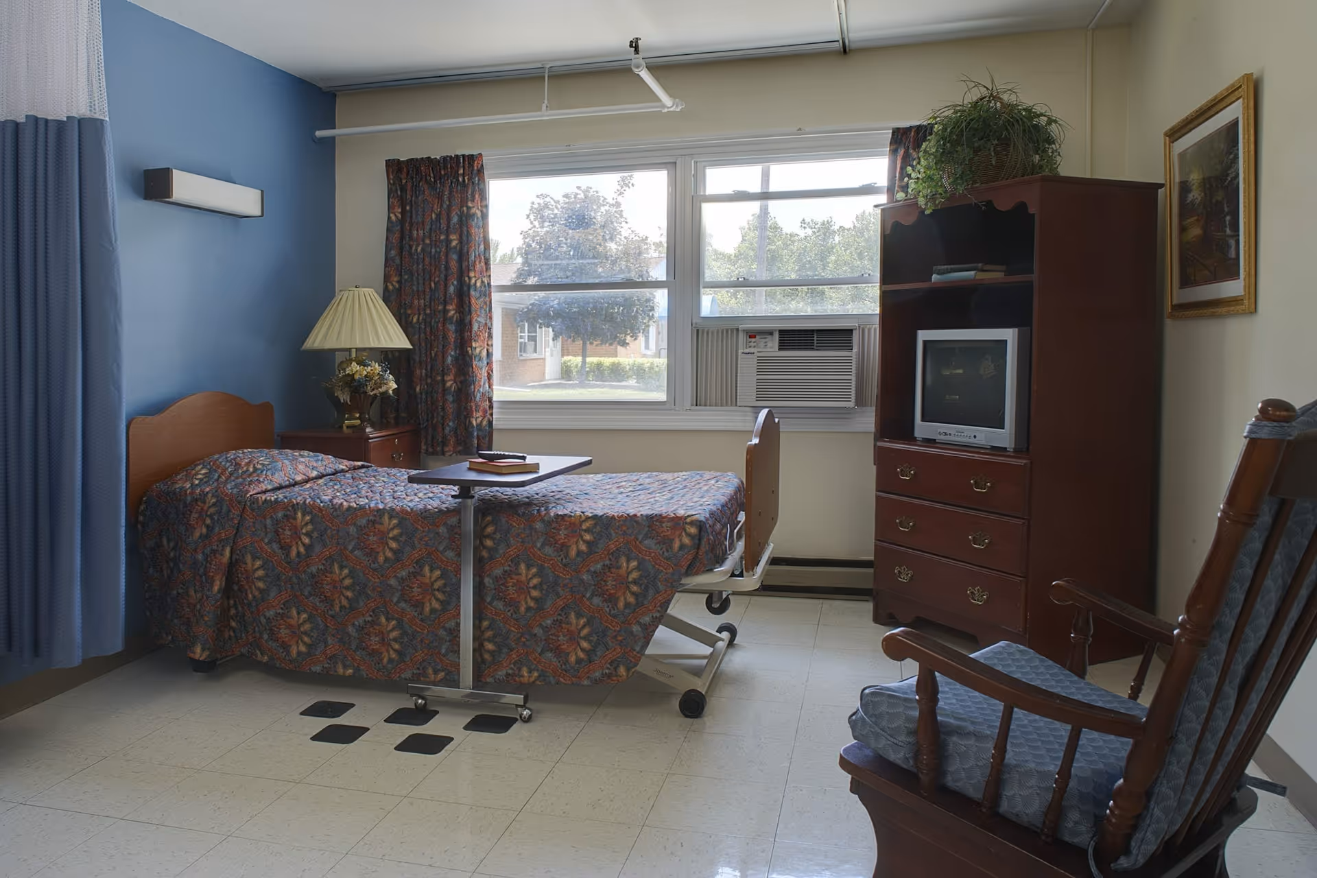 A simple senior living bedroom with a single bed covered in a patterned bedspread, a bedside table with a lamp and flowers, a rolling overbed table with a book and remote control, a wooden rocking chair with a cushion, a wooden dresser with a small TV and a potted plant on top, and a window with floral curtains and an air conditioning unit.