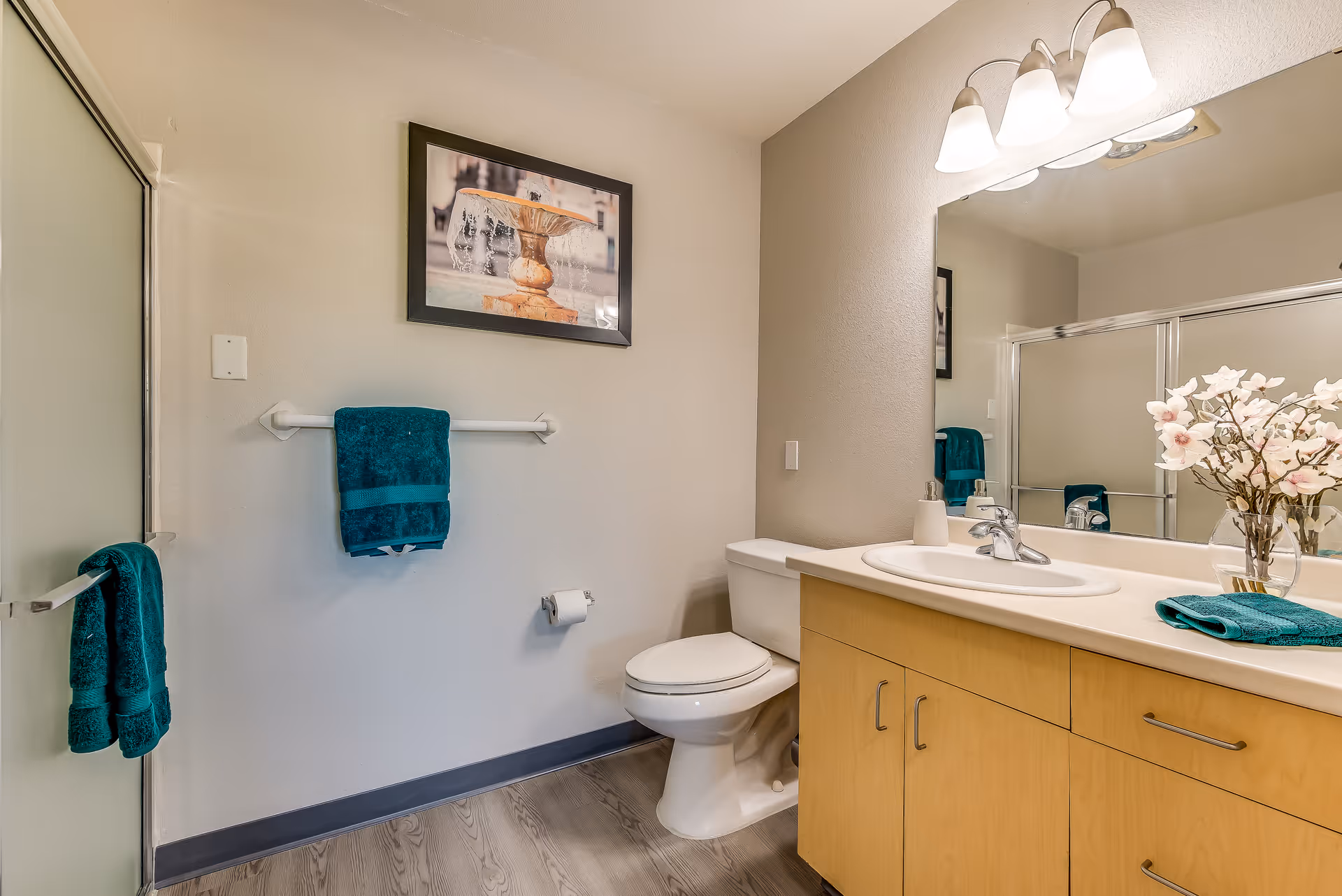 A clean bathroom with a wooden vanity featuring a white sink and silver faucet. Above the vanity is a large mirror with three light fixtures. To the right of the vanity is a white toilet. On the wall above the toilet is a framed picture of a fountain. There are teal towels hanging on a towel rack and a hook on the left wall. The floor has a wood-like finish, and a glass shower door is partially visible on the left side.