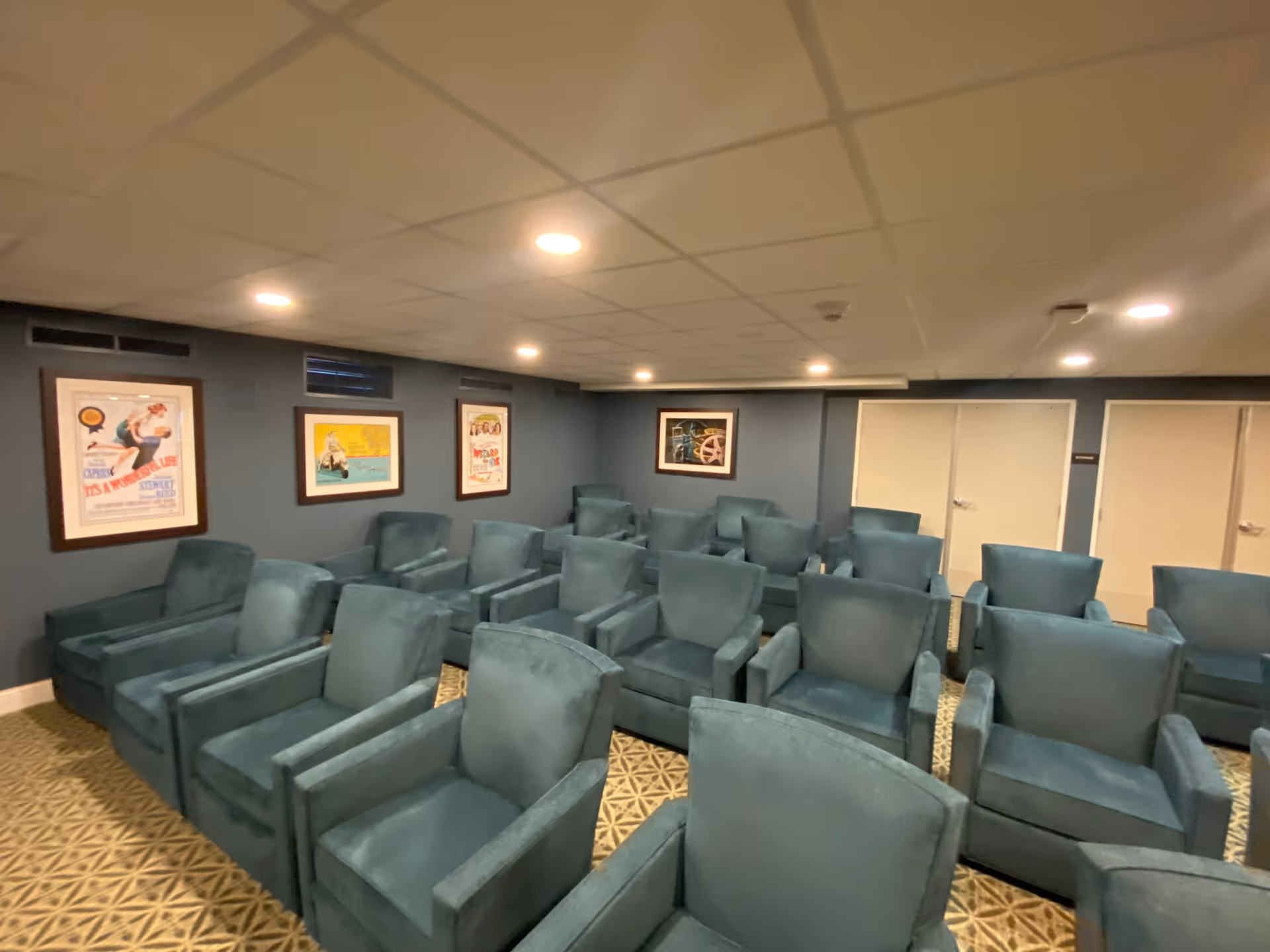 A small theater or presentation room with multiple rows of teal upholstered armchairs arranged facing forward. The walls are painted blue and decorated with framed vintage-style posters. The ceiling has recessed lighting and there are two closed white doors at the back of the room.