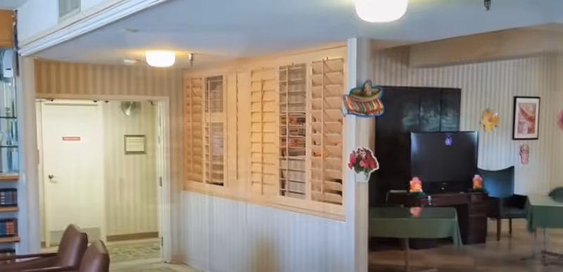 Interior view of a senior living facility showing a common area with green tables and chairs, a television on a dark wooden stand, and decorative wall hangings including flowers and a sombrero. There is a window with wooden shutters and a doorway leading to another room with a white door.
