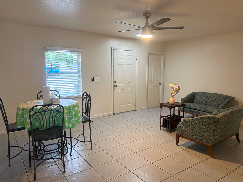 Small living room and dining area with tiled floor, a round table with a green floral tablecloth and chairs, two upholstered sofas, a small side table with a vase, and a ceiling fan.