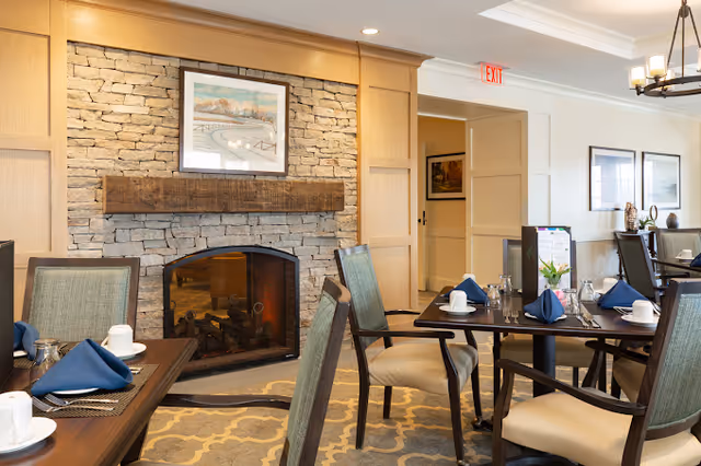 A cozy dining room with several tables set with white cups, plates, silverware, and blue napkins. The room features a stone fireplace with a wooden mantel and a framed painting above it. The walls are light-colored with wooden paneling, and there are framed pictures on the far wall. The room has a warm and inviting atmosphere.