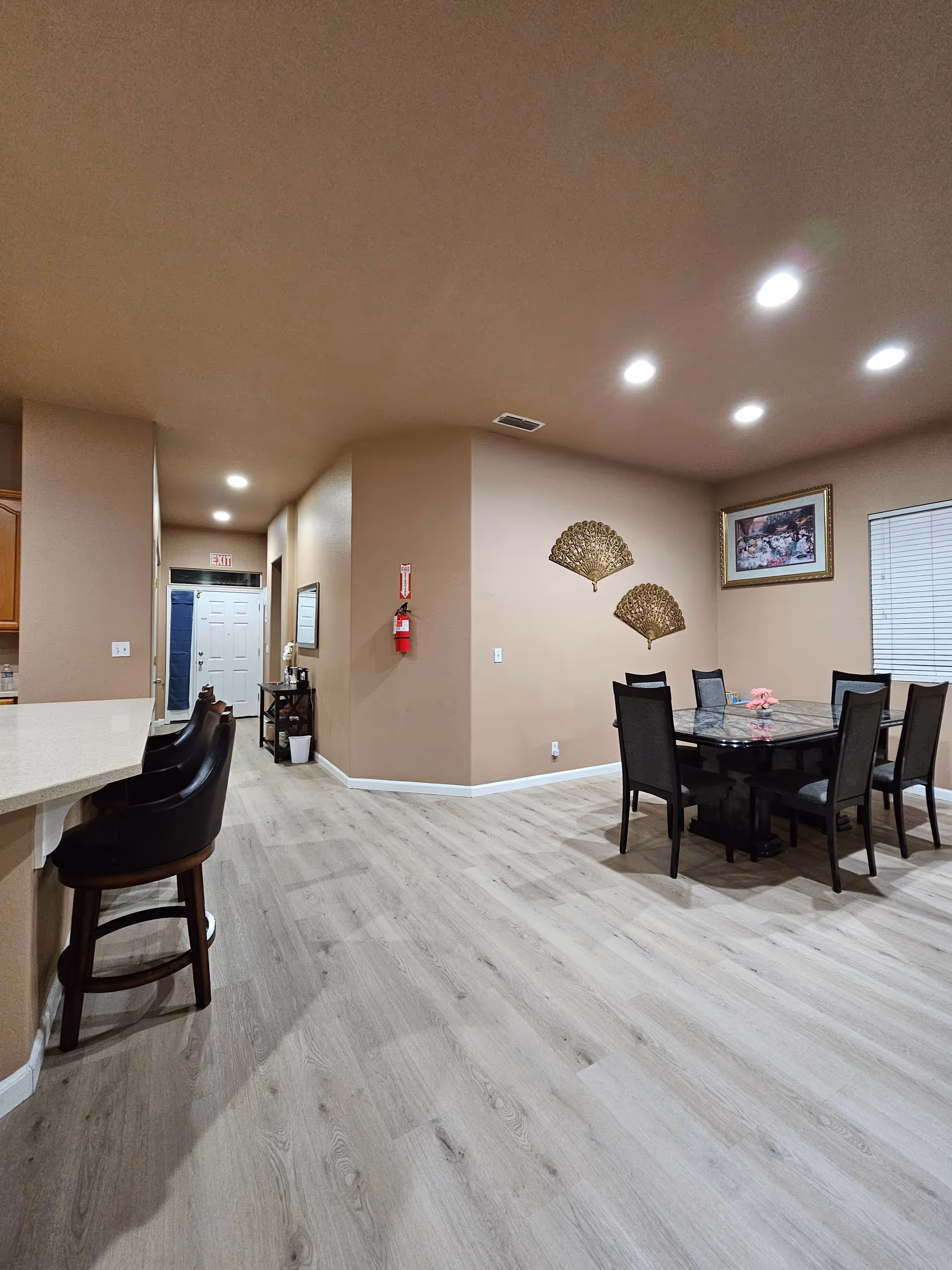 Open interior dining area with a dining table and chairs, bar stools at a counter, wall decor, and a hallway with an exit door.