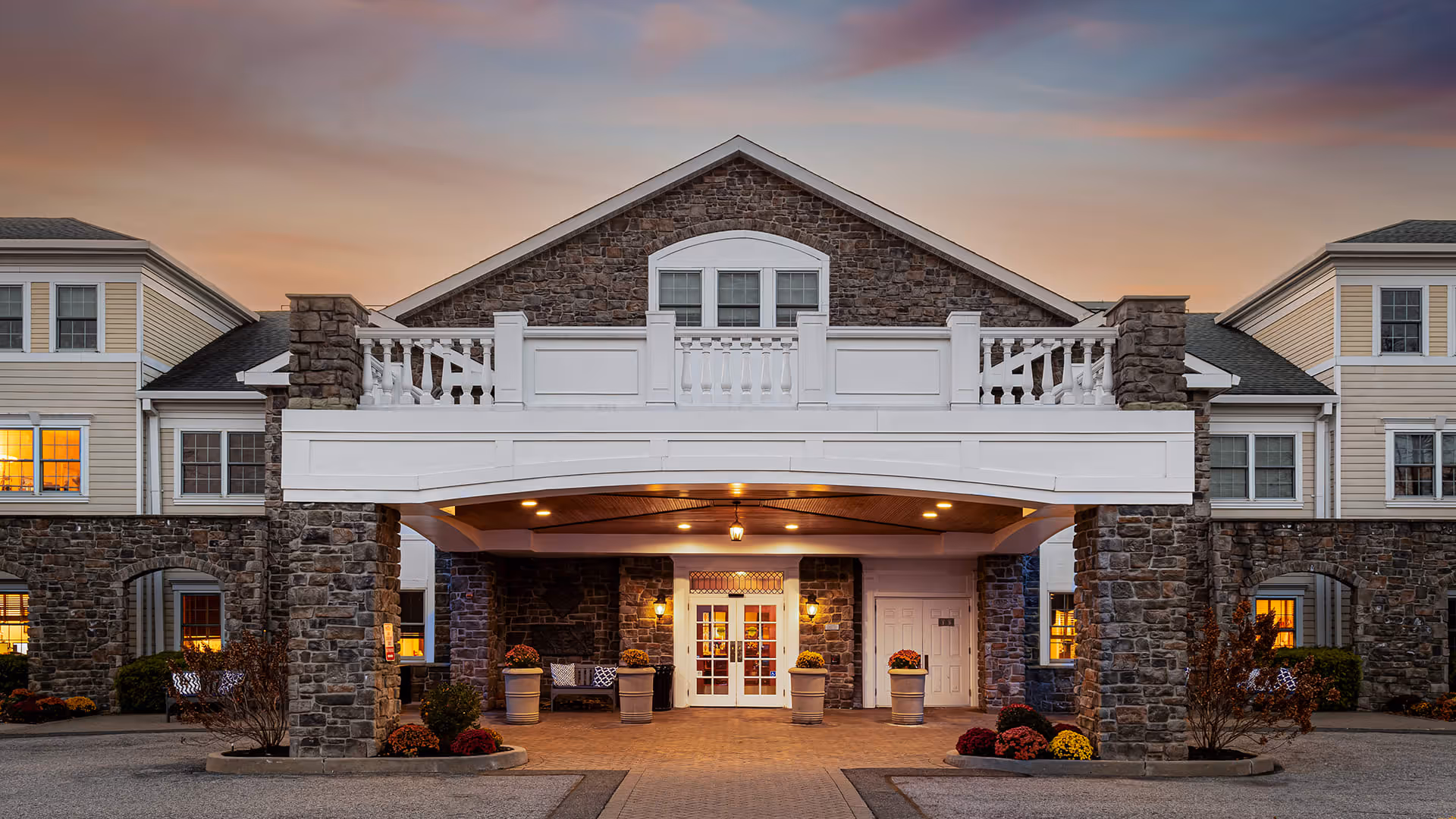 Front exterior view of Atria Rye Brook facility at dusk, featuring a stone and siding building with a covered entrance supported by stone pillars, illuminated by warm lights, and landscaped with potted plants and flowers.