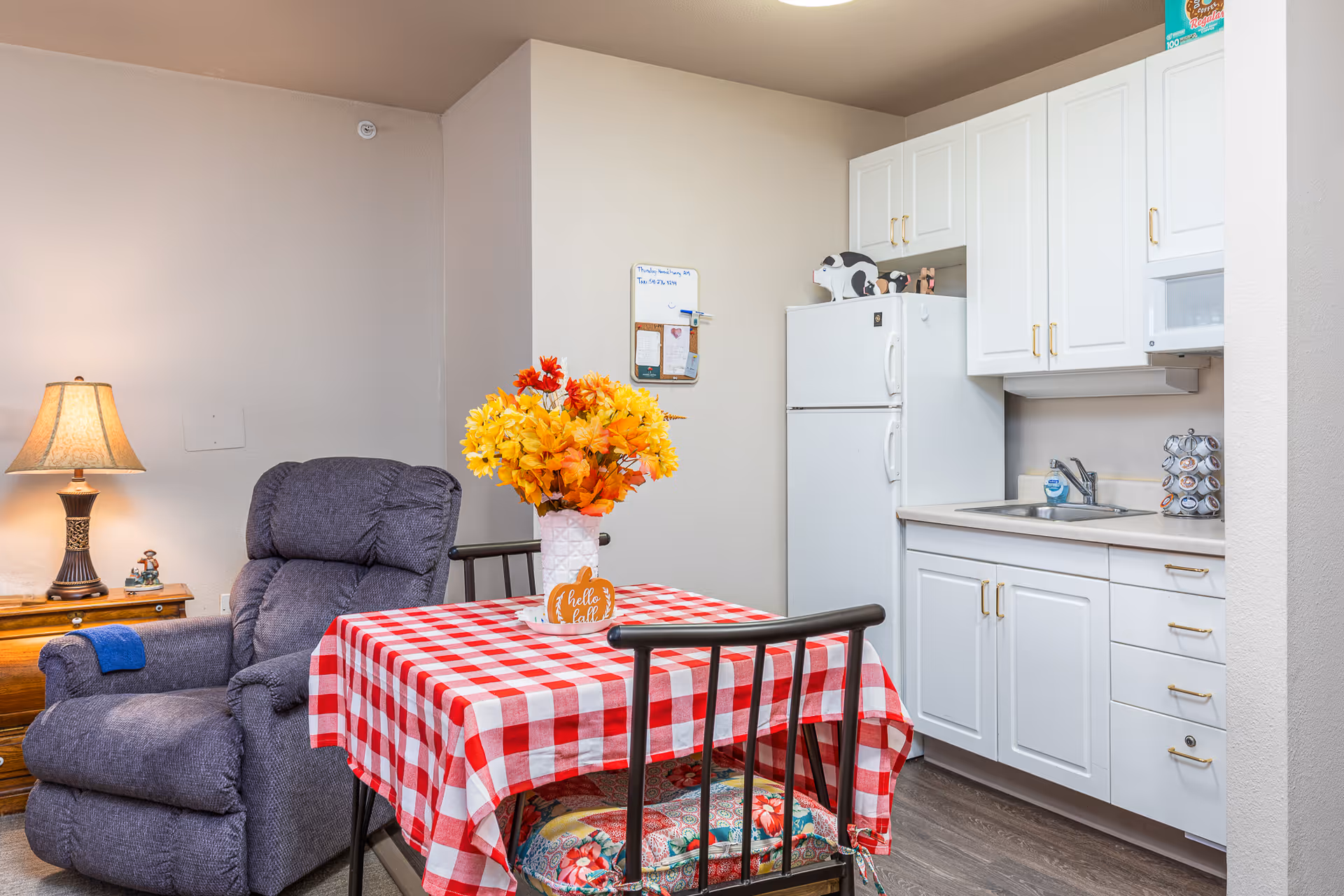 A cozy assisted living kitchen and dining area featuring white cabinets, a white refrigerator with decorative items on top, a small sink, and a microwave. In front of the kitchen is a table covered with a red and white checkered tablecloth, adorned with a vase of bright yellow and orange flowers. Next to the table is a comfortable dark gray recliner chair with a small wooden side table and a lamp beside it.
