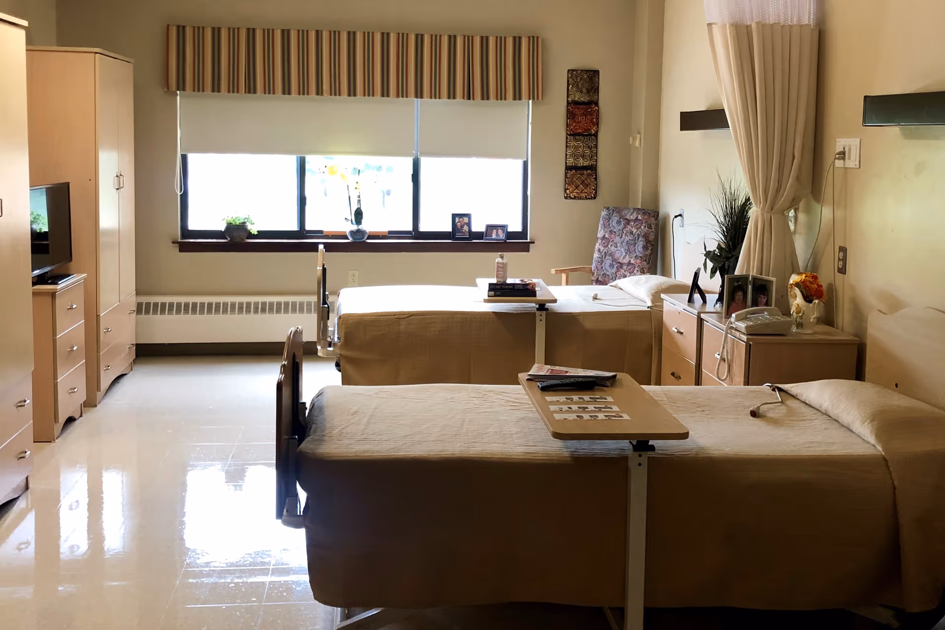 A well-lit room with two hospital-style beds covered in beige bedspreads. Each bed has an overbed table with items on top, such as a remote control and books. There is a window with blinds partially drawn, a floral-patterned armchair, a small dresser with framed photos, a telephone, and a TV on a cabinet. The room has a clean, simple, and functional design.