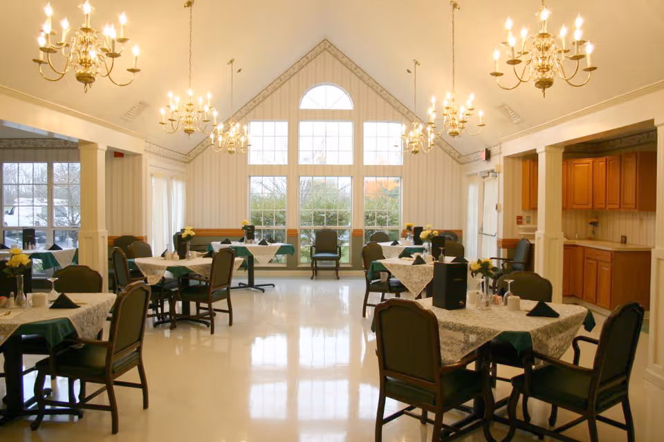 Bright dining room with multiple tables set with green tablecloths and chandeliers and large arched windows at the far wall.