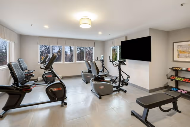 Bright exercise room with several stationary bikes, a bench, free weights on shelves, and a wall-mounted TV.