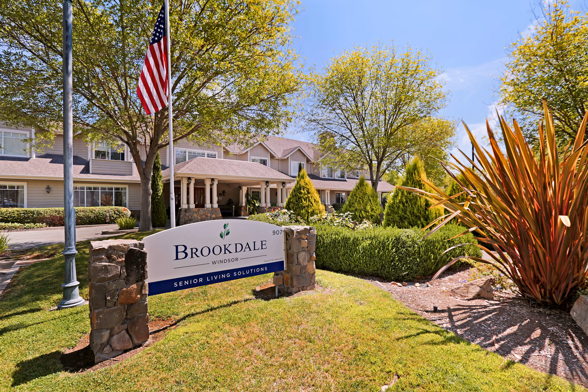 Exterior view of Brookdale Windsor senior living facility with a large sign in the foreground, an American flag on a flagpole, well-maintained landscaping including trees and shrubs, and a two-story building in the background under a clear blue sky.