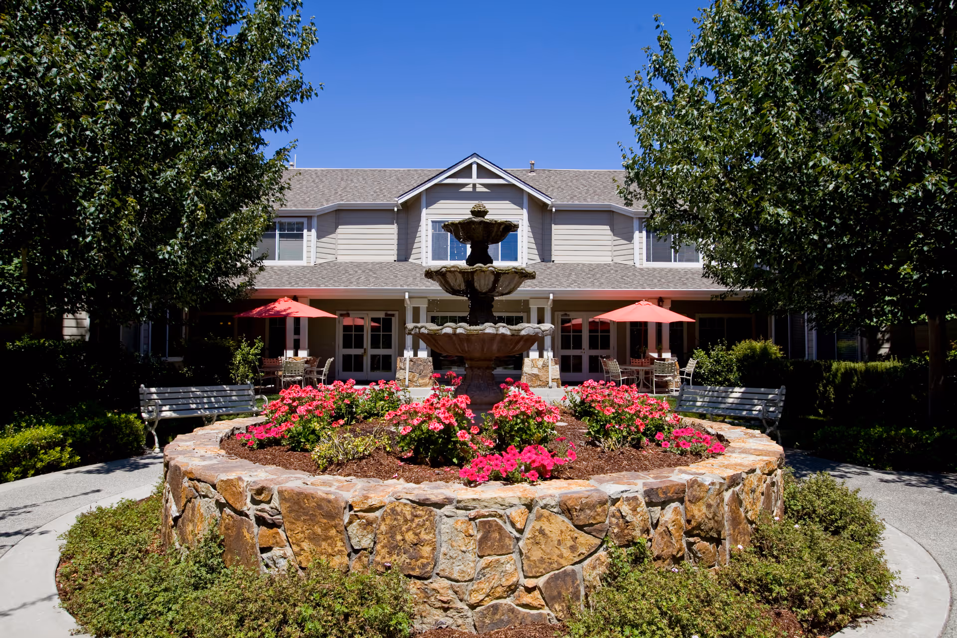 Outdoor courtyard area of a senior living facility with a stone circular planter filled with pink flowers and a three-tiered water fountain in the center. There are benches on either side of the planter and red patio umbrellas shading tables and chairs in front of a two-story building with beige siding and white trim. Trees frame the courtyard under a clear blue sky.