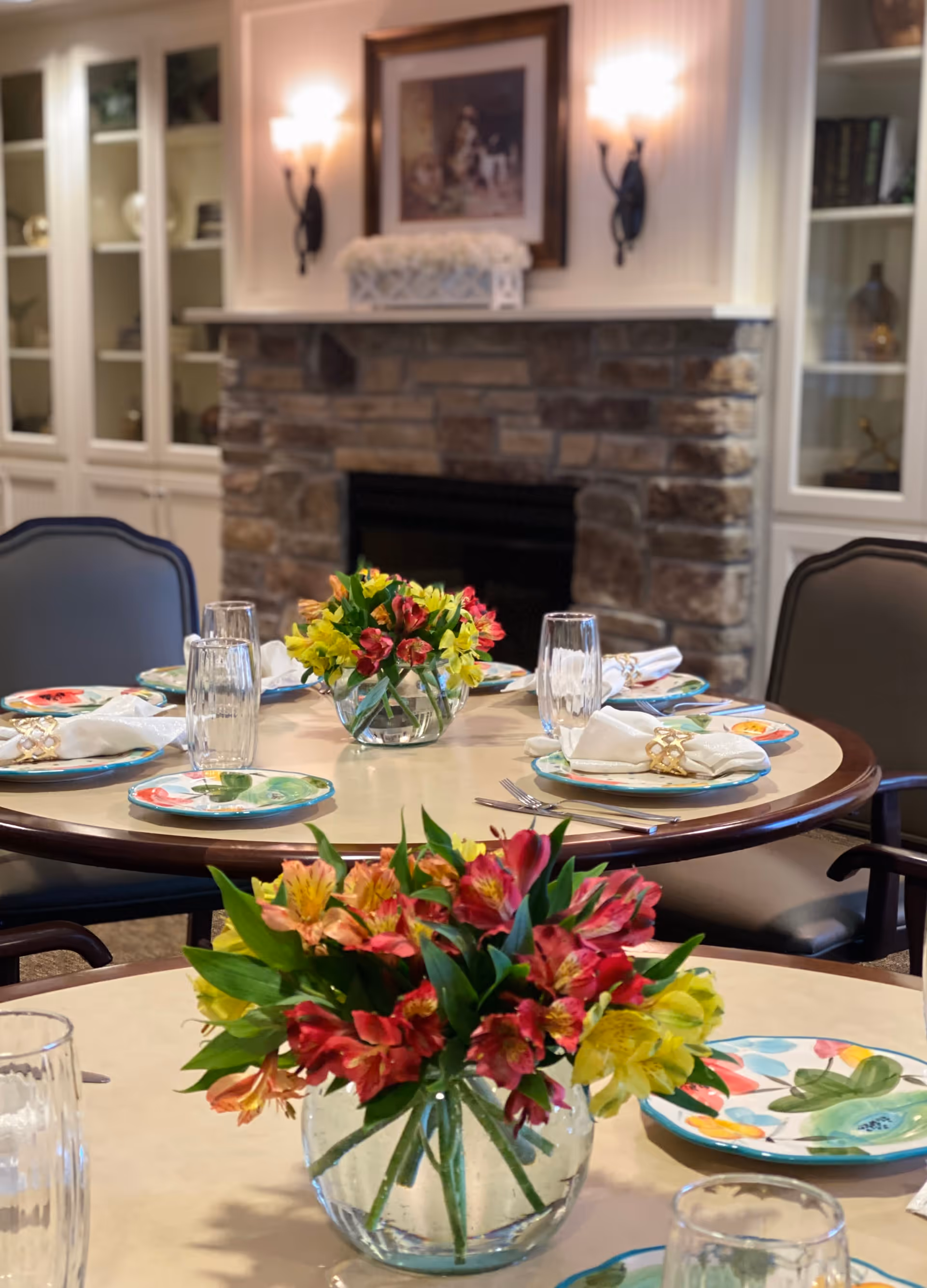 A cozy dining area with round tables set for a meal, featuring colorful floral plates, glassware, and napkins with napkin rings. Each table has a clear glass vase with vibrant red, yellow, and orange flowers. In the background, there is a stone fireplace with a framed picture above it and built-in white shelves on either side.