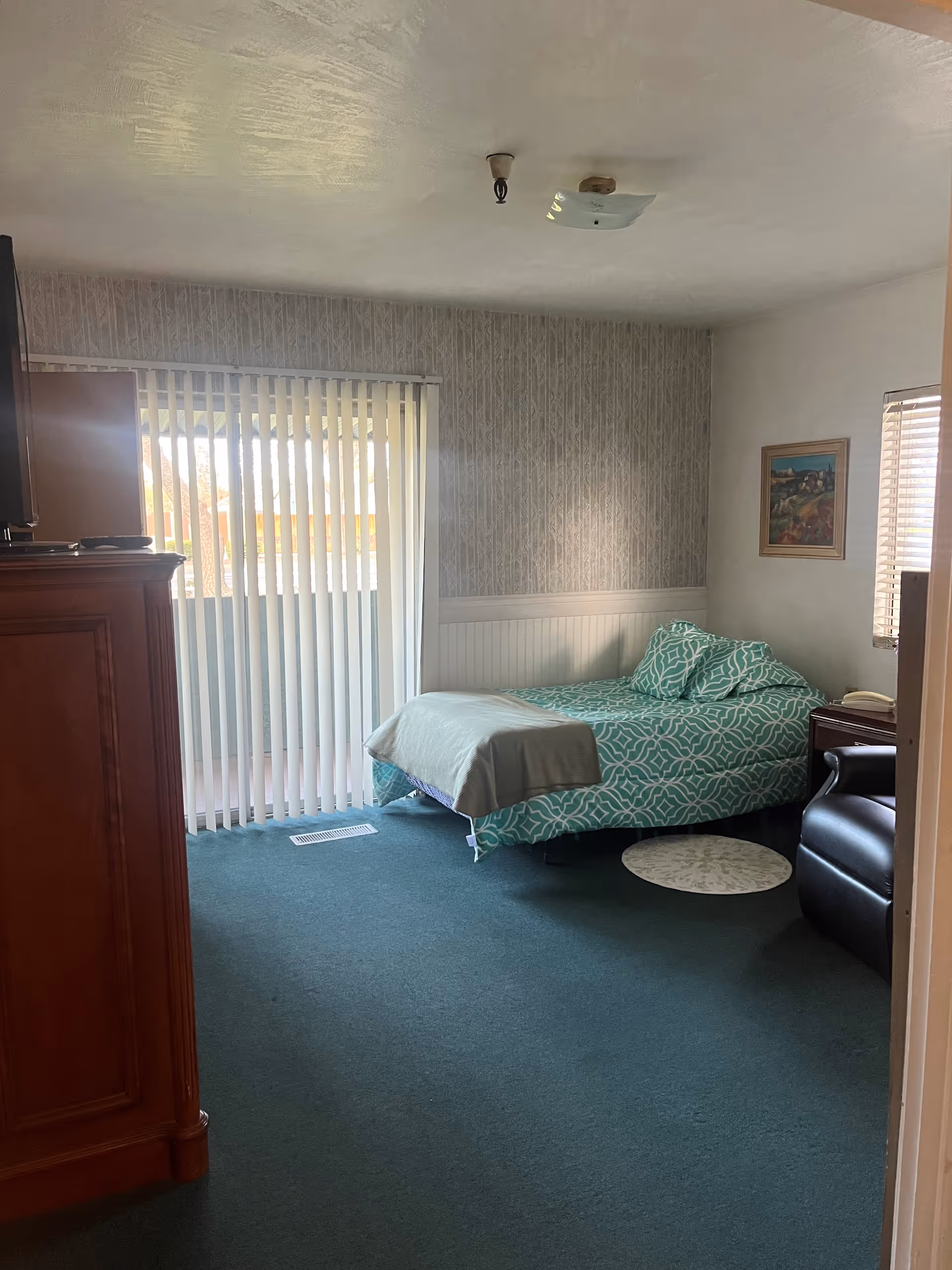 A senior living facility bedroom with a single bed covered in a teal and white patterned bedspread and matching pillows. The room has green carpet, a wooden dresser, a black recliner chair, a small round rug, and a framed painting on the wall. Vertical blinds cover a sliding glass door leading to an outdoor balcony or patio.