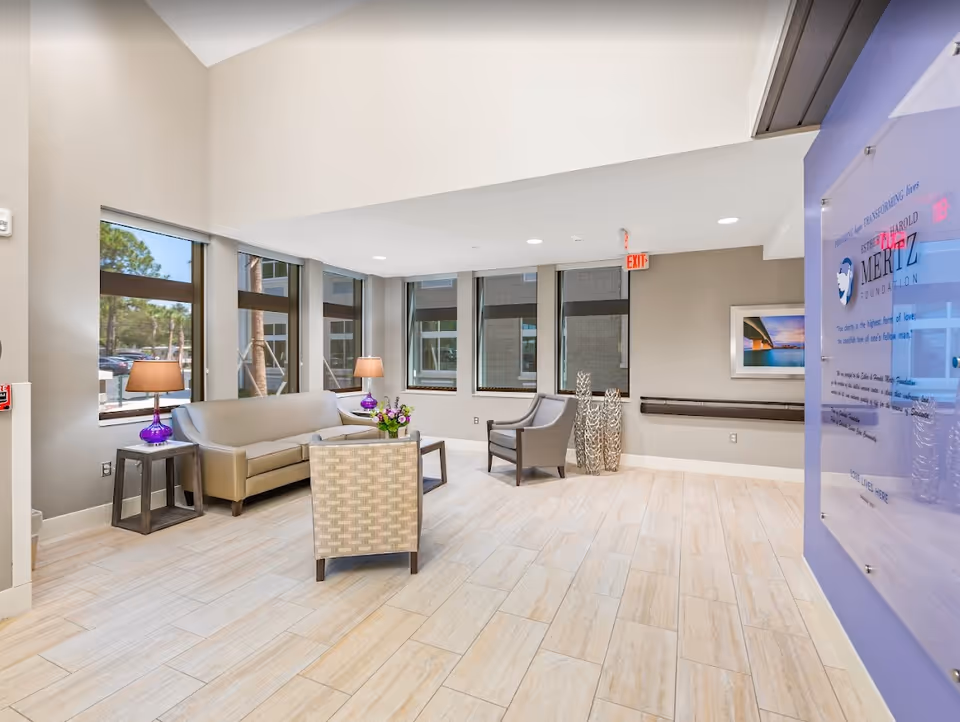 A bright and modern lounge area with beige tiled flooring, a beige sofa, two armchairs, two side tables with purple lamps, and a coffee table with a flower arrangement. Large windows let in natural light, and a blue informational board is mounted on the right wall. The space has a high ceiling and neutral-colored walls.