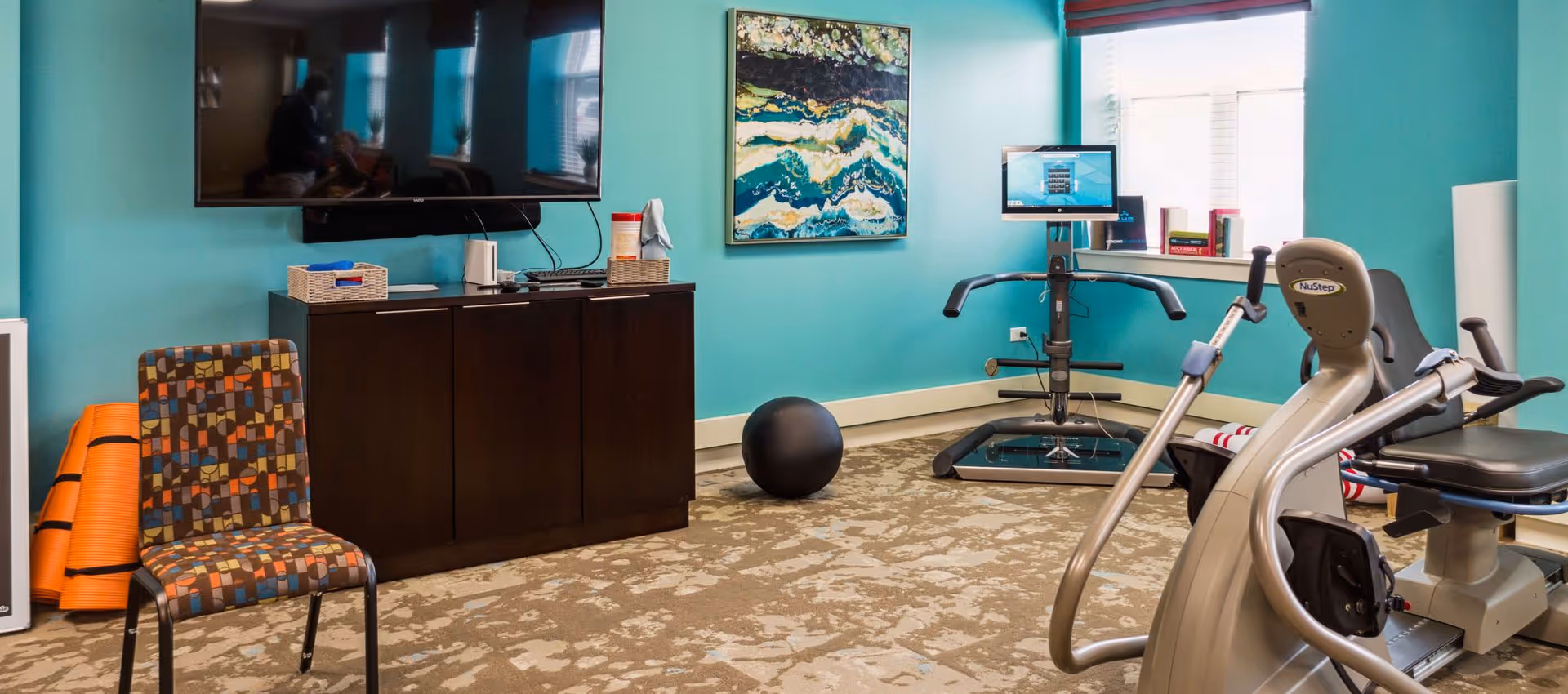 A small exercise room with teal walls featuring a recumbent exercise bike, a NuStep machine, a black exercise ball, a patterned chair, a dark wooden cabinet with a large flat-screen TV mounted above it, and a colorful abstract painting on the wall. There is a window with blinds letting in natural light.