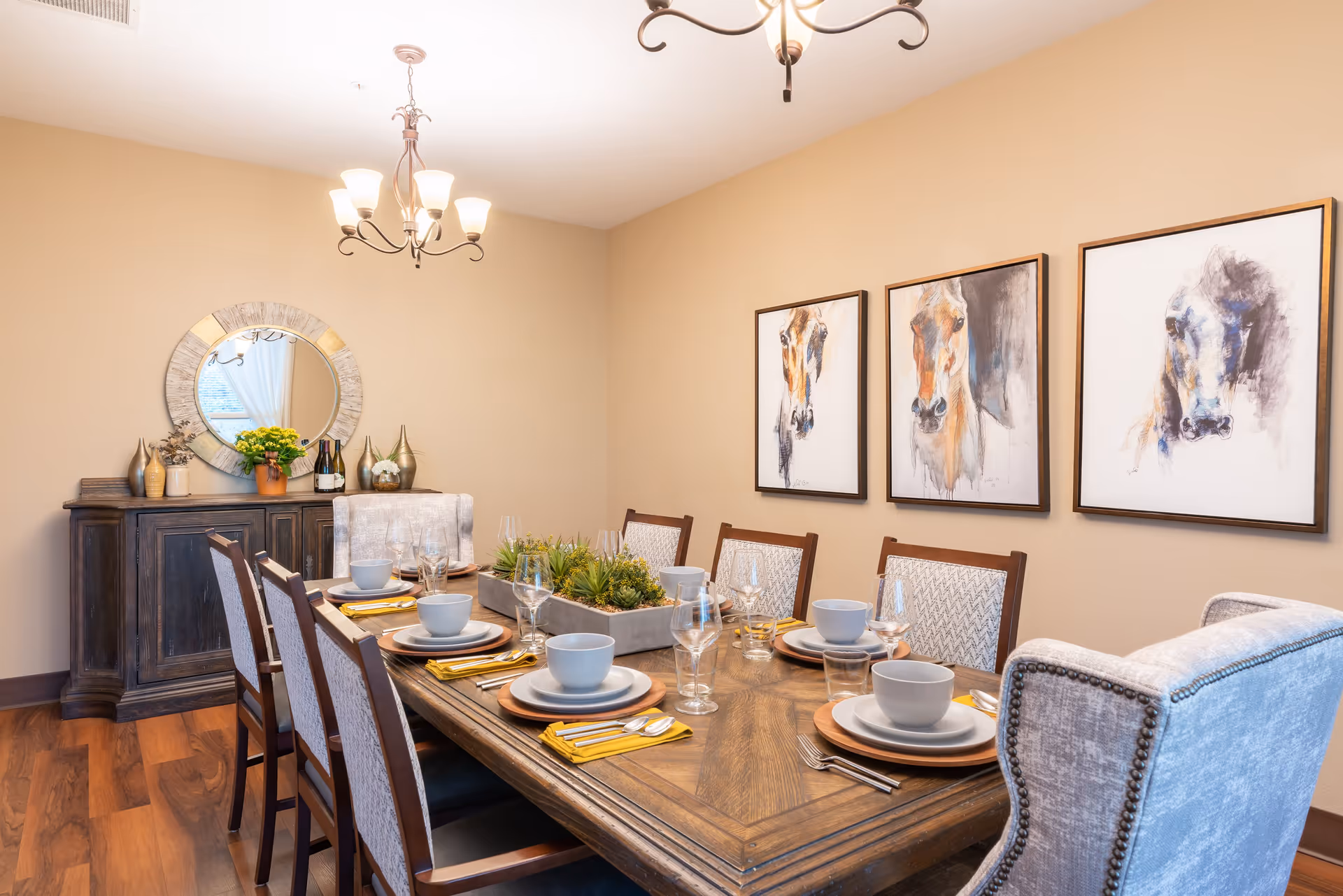 A dining room with a wooden table set for six people, featuring plates, bowls, glasses, and yellow napkins. The room has wooden flooring, beige walls, and three framed paintings of horses on one wall. A dark wooden sideboard with decorative items and a round mirror is against another wall. Two ceiling light fixtures hang above the table.