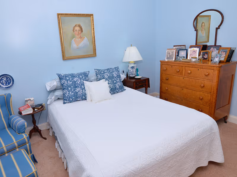 Bedroom with a white bedspread and blue pillows, light blue walls, a wooden dresser topped with framed photos, bedside tables, and a striped armchair.