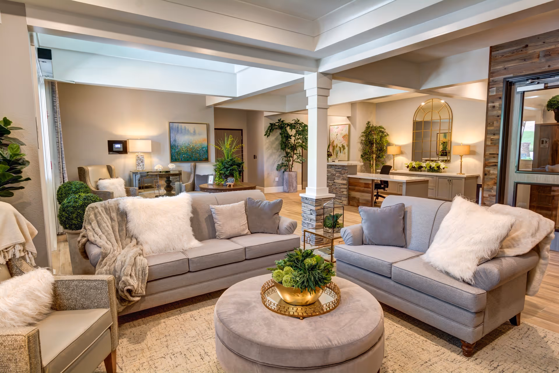 A cozy and well-lit living room area with two gray sofas adorned with white fluffy pillows and a gray throw blanket. A round ottoman with a decorative plant in a golden pot sits in the center on a patterned rug. The room features wooden flooring, potted plants, a desk with a chair, lamps, and framed artwork on the walls, creating a warm and inviting atmosphere.