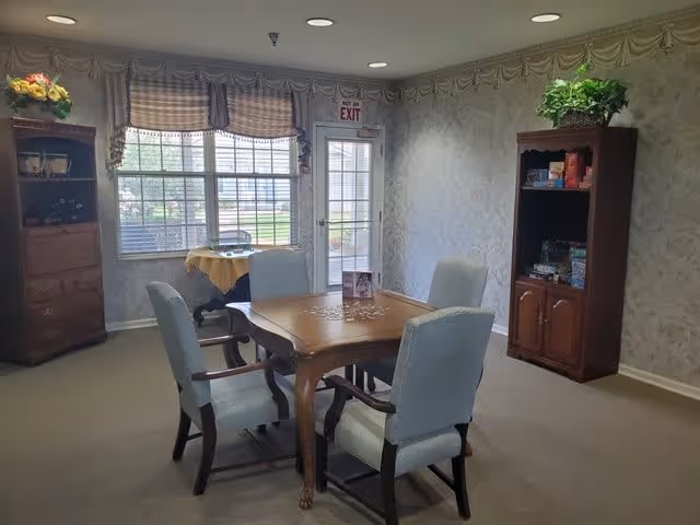 A cozy room with a wooden table surrounded by four upholstered chairs. The room has patterned wallpaper, a window with plaid valances, and a glass door with an exit sign above it. There are two wooden cabinets, one with board games and a plant on top, and the other with decorative items and flowers.