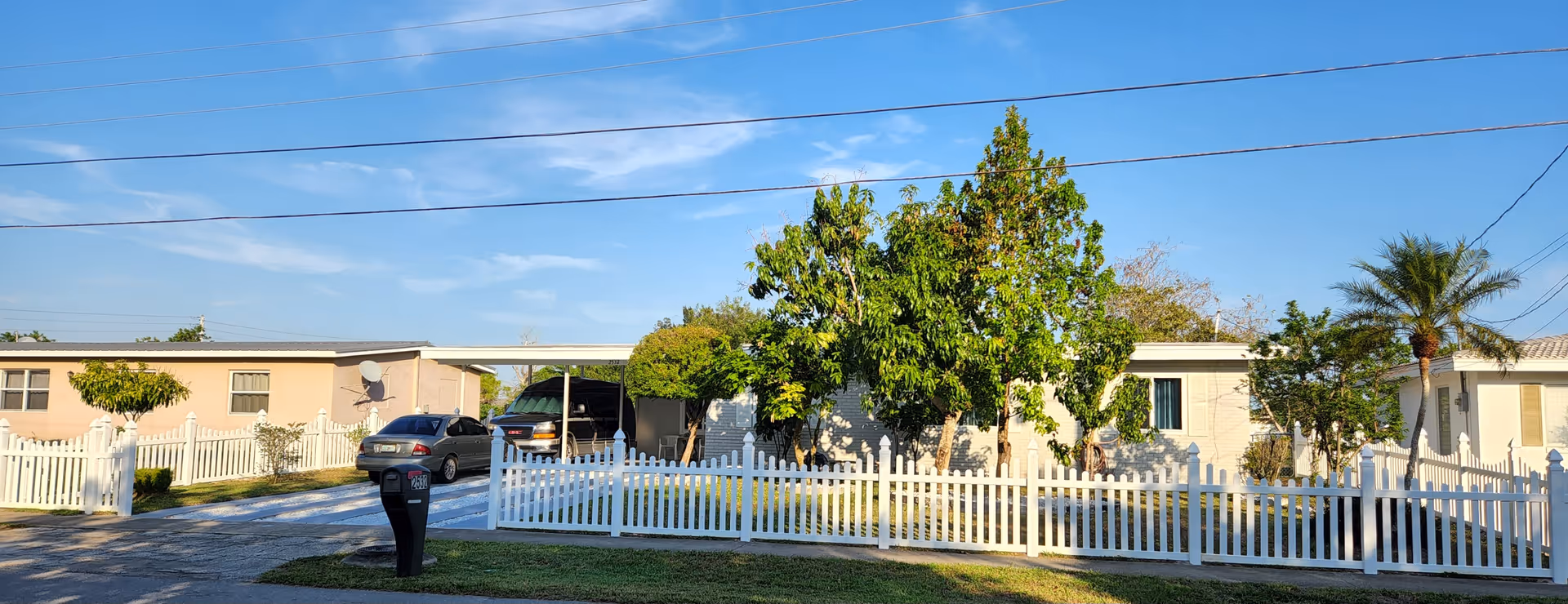 Single-story residential units behind a white picket fence with parked cars and trees under a blue sky.