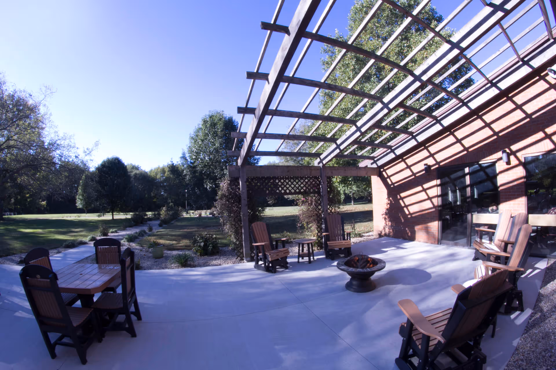 Outdoor patio area with wooden chairs and tables under a pergola attached to a brick building. The patio overlooks a grassy area with trees and a clear blue sky.