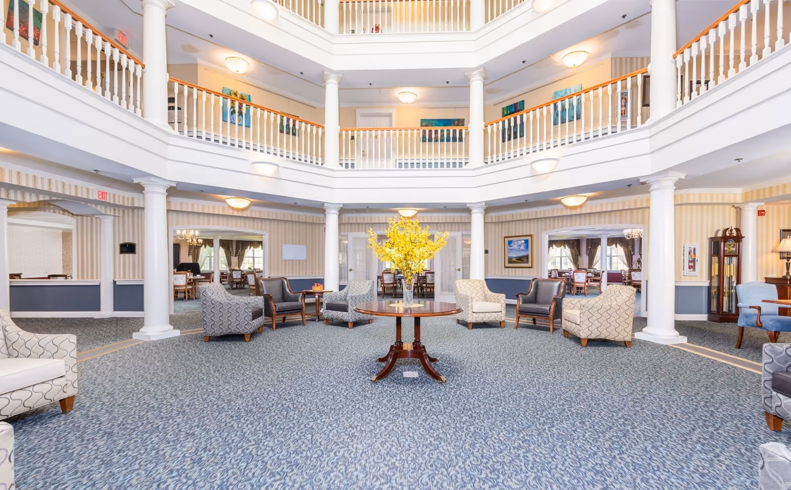 Spacious, well-lit common area in a senior living facility with a round wooden table holding a vase of yellow flowers in the center. Surrounding the table are multiple upholstered armchairs arranged in a circle. The room features white columns, a blue patterned carpet, and a two-story balcony with white railings. The walls have light-colored striped wallpaper and framed artwork. Adjacent rooms with dining tables and chairs are visible through large open doorways.
