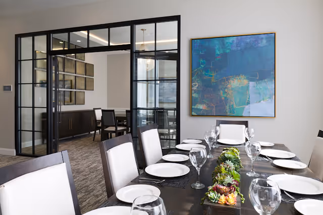 A modern dining room with a long dark wooden table set with white plates, wine glasses, and a succulent centerpiece. The room features white cushioned chairs and a large abstract painting on the wall. Glass and black metal framed double doors open to an adjacent room with additional seating and wall art.